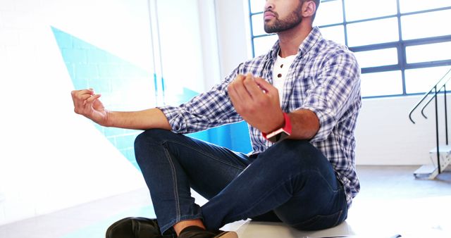 This image shows a young man in casual attire practicing meditation in an office environment. Ideal for promoting mental health, workplace wellness, stress-relief activities, mindfulness practices, employee productivity, and self-care. Useful for blogs, corporate wellness programs, stress management workshops, and health-related articles.
