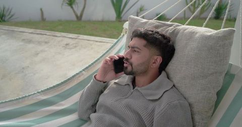 Indian man relaxing in striped hammock on patio talking on smartphone for backyard comfort
