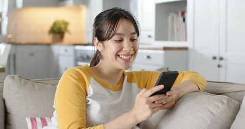 Smiling asian woman relaxing on sofa using smartphone
