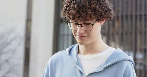 Young man wearing round glasses and blue hoodie smiling looking down in urban portrait