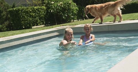 Mother and daughter playing in backyard pool with golden retriever on pool deck
