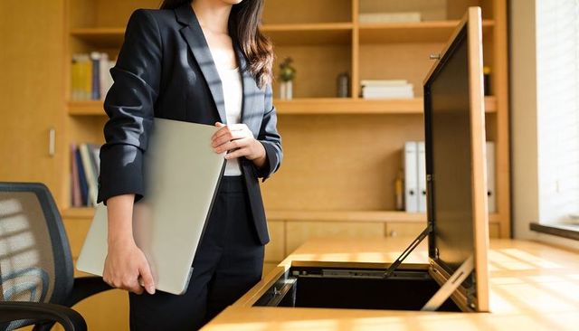 Asian businesswoman holding laptop beside open desk compartment in sunlit wooden office