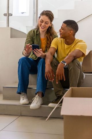 Young couple sitting on stairs unpacking moving boxes and checking smartphone in new home