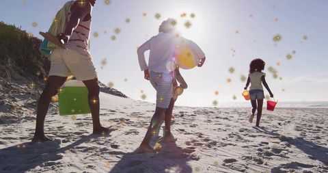 Family walking toward ocean on sunlit beach carrying cooler, board, ring and sand buckets
