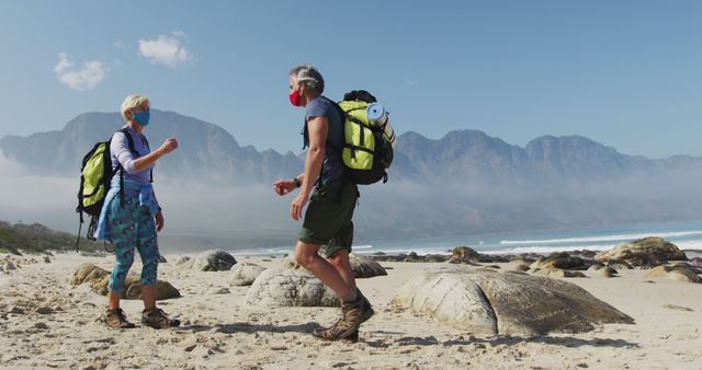 Senior hiker couple wearing face masks with backpacks greeting each other by touching elbows while hiking on the beach. trekking, hiking, nature, activity, exploration, adventure concept.