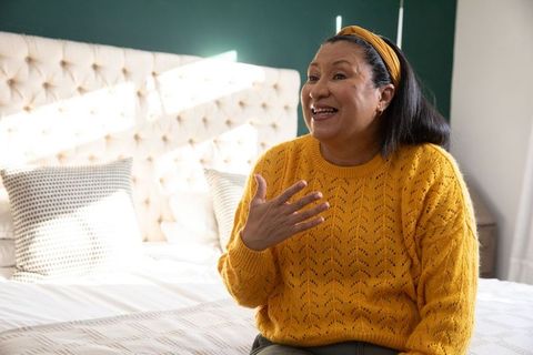 Smiling woman sitting on bed wearing mustard knit sweater and headband, cozy bedroom moment