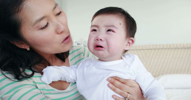 An Asian mother comforts her crying baby, with copy space. Her gentle expression suggests concern and care as she tries to soothe her child's distress.