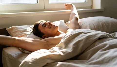 Young man stretching in morning sunlight on minimalist bed, relaxed wakeup