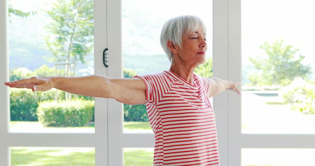 Senior woman wearing striped shirt practicing yoga with arms outstretched in bright room filled with natural light. Ideal for depicting themes of wellness, fitness for seniors, healthy aging, mindfulness, and everyday tranquility. Useful for blogs, articles, and advertisements focusing on senior health, fitness programs, and mental well-being.
