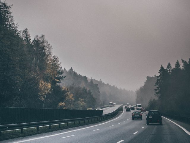 Cars driving along a foggy highway bordered by dense forest during autumn. The trees display muted colors due to the fog, creating a calm and somewhat mysterious atmosphere. It is suitable for use in projects related to travel, road trips, seasonal changes, and transportation.