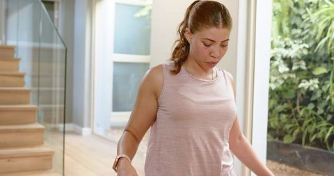 Young woman practicing balance indoors near glass door and wooden stairs with smartwatch