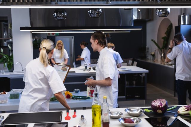 Group of chefs cooking in a modern busy kitchen, female chef holding a clipboard giving instructions. Cookery class at a restaurant kitchen. Workshop cooking food.