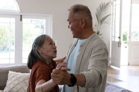 Senior asian couple dancing slowly together in sunlit living room showing affection and joy