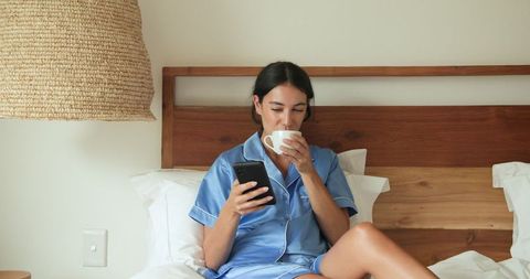 Woman Relaxing on Bed in Cozy Satin Pajamas with Smartphone