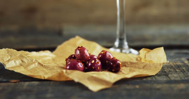 Candied nuts on crumpled paper on rustic wooden surface. Blurred glass in background adds elegance. Ideal for food blogs, recipe websites, gourmet snack promotions, restaurant menus, or rustic kitchen decor.