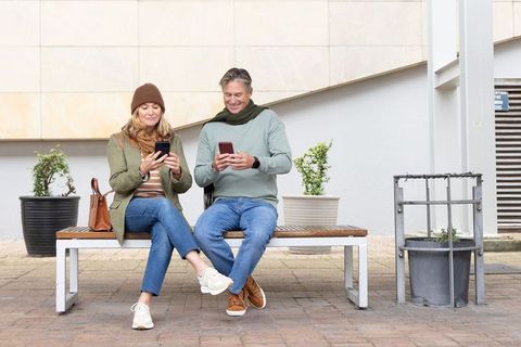 Middle-aged couple sitting on bench scrolling smartphones in modern urban courtyard
