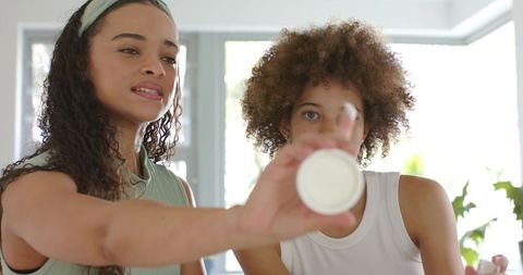 African american women demonstrating natural skincare with cream jar in sunlit home beauty
