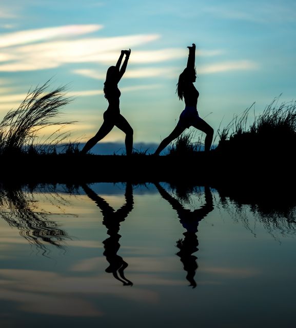 Two women are practicing yoga and stretching poses near a serene lake at sunset. Their silhouettes are reflected in the calm water, creating a tranquil and peaceful scene. This can be used for promoting outdoor fitness, yoga retreats, health and wellness campaigns, nature-inspired relaxation methods, or meditation exercises.