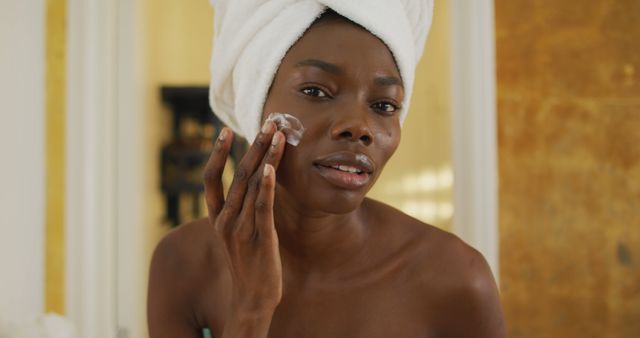 Portrait of smiling african american woman with towel using cream on her face in bathroom. health and beauty concept.