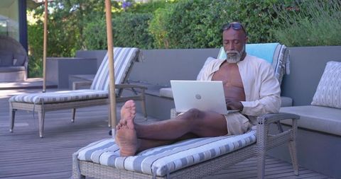 Senior african man lounging on patio chaise using laptop for remote work and relaxation