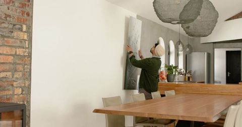 Indian man hanging contemporary art panel in open-plan dining kitchen with timber table