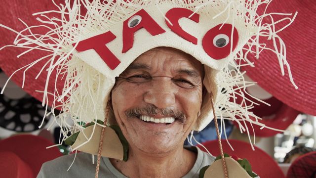 A senior man displays his bright smile while wearing a playful hat adorned with humorous embellishments. The lively colors and various hats displayed in the backdrop create an engaging and cheerful atmosphere, representing joy and creativity in self-expression. Useful for themes related to aging with positivity, humor in daily life, and creative showcases in a casual, whimsical setting. Suitable for marketing purposes, social media discussion on aging or lifestyle articles promoting joyful living.