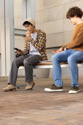 Teen friends eating sandwich and checking phone on campus bench, casual student life