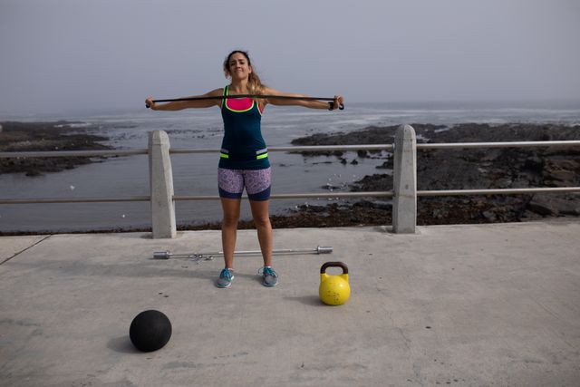 Front view of a strong Caucasian woman with long dark hair wearing sportswear exercising outdoors by the seaside on a sunny day, strength training stretching black rubber tape with her arms, ball and kettlebell next to her.