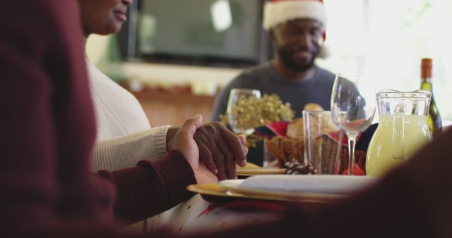 African american family in santa hats holding hands and praying together sitting on dining table before having lunch at home.