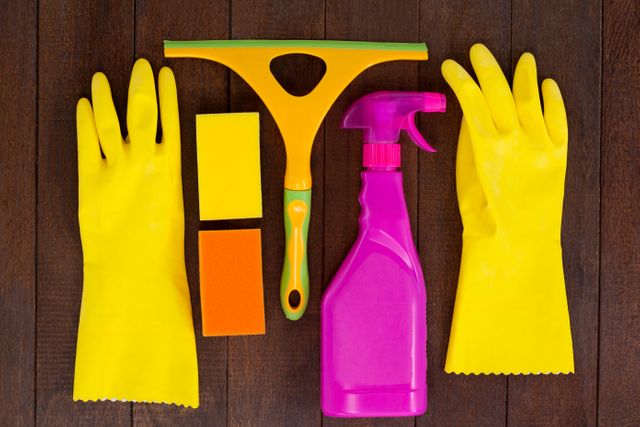 Cleaning supplies including rubber gloves, a spray bottle, sponges, and a squeegee are neatly arranged on a wooden floor. This image is ideal for illustrating articles, advertisements, or blog posts related to housekeeping, cleaning tips, and home maintenance. It can also be used in promotional materials for cleaning products or services.