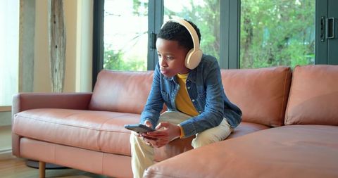 African american boy wearing headphones sitting on tan sofa holding smartphone in living room