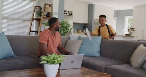 Work-from-home father using laptop while son handing homework on living room couch