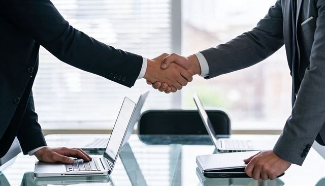 Business leaders shaking hands over glass conference table with laptops reflecting