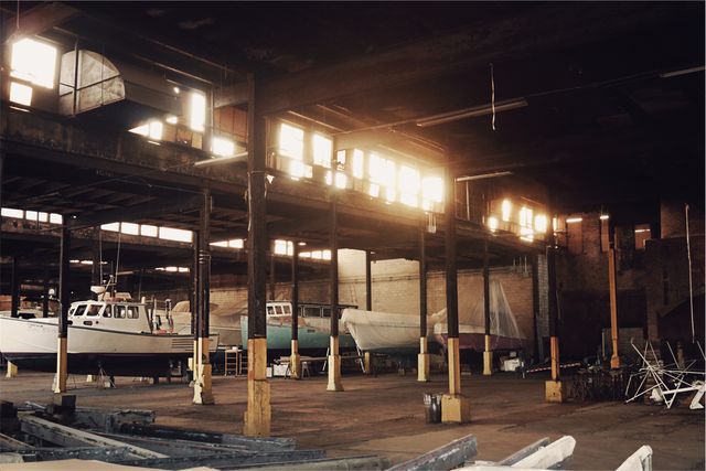 This image showcases the interior of an industrial warehouse with several docked boats bathed in warm sunlight filtering from the upper windows. The large, spacious area with rustic beams and support structures creates a unique atmosphere. Ideal for usage in topics related to industry, nautical themes, boat storage, and rustic architecture. Perfect for blogs, articles, and websites interested in industrial design and maritime environments.