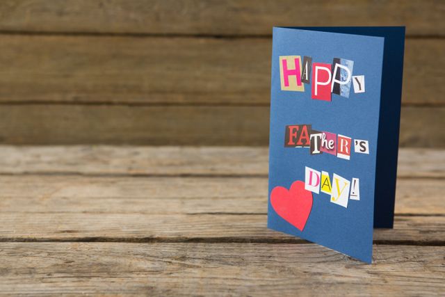 Handmade Father's Day card with colorful cut-out letters and a red heart on a blue card, placed on a rustic wooden table. Ideal for use in holiday promotions, DIY craft tutorials, and Father's Day celebration advertisements.