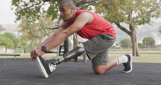 Man with a prosthetic leg is stretching his legs while wearing athletic attire in an outdoor gym area, surrounded by trees. This image can be used to promote fitness, inclusivity, and the inspirational stories of individuals overcoming physical challenges. Suitable for articles on sports, health, motivational campaigns, and disability awareness.