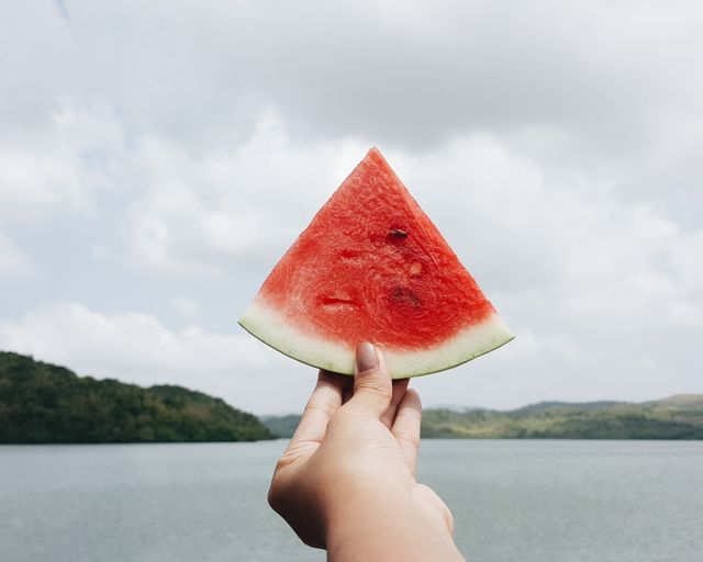 Person holding slice of watermelon outdoors near lake on cloudy day. Great for summer vacation promotions, refreshing snacks, outdoor activities, and health and wellness concepts.