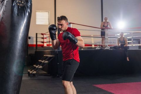 Male boxer training with heavy bag in gym ring spotlighted, focused stance