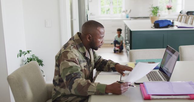 A military father in camouflage uniform working from home on a laptop, holding documents while a child plays in the background. Ideal for themes such as work-life balance, military families, remote work, home offices, and parenting dynamics.
