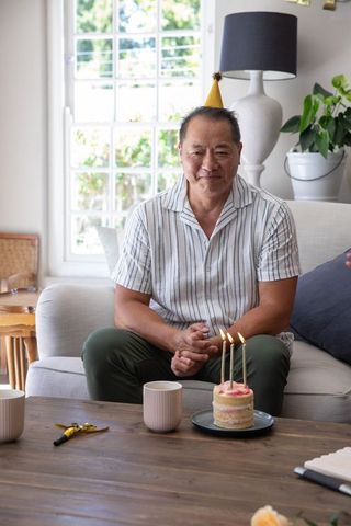 Middle-aged chinese man sitting on sofa celebrating birthday with cake and candles at home