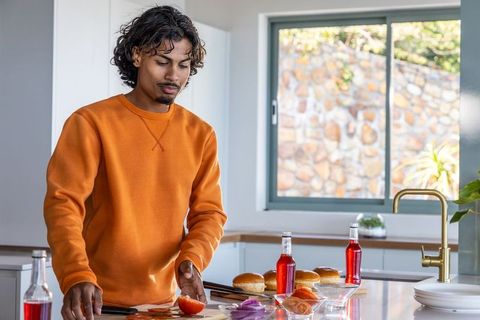 Indian young man slicing tomato and arranging burger buns on modern kitchen island