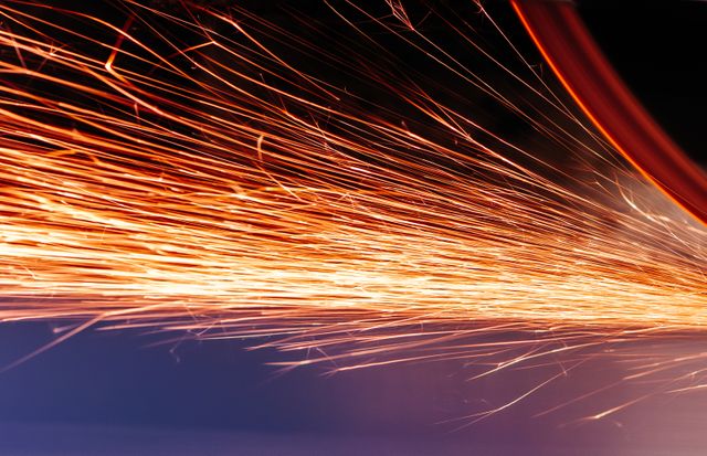 Sparks flying off a high-speed industrial grinder, creating a dynamic and energetic scene. Ideal for depicting metalworking processes, industrial craftsmanship, manufacturing environments, and scenes that require a sense of motion and activity. Useful for websites, brochures, or articles on engineering, manufacturing, and mechanical workshops.