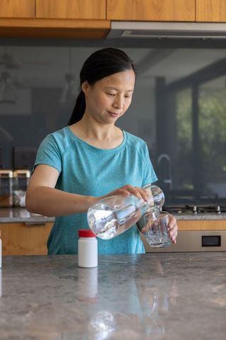 Mature asian woman pouring water into glass in modern sunlit kitchen promoting hydration