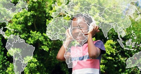 Smiling boy listening to headphones with world map overlay in sunlit green park