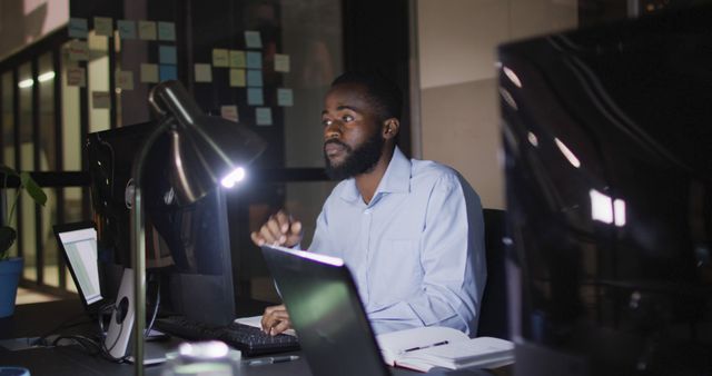 African American man working late in modern office, surrounded by computers and office equipment. Ideal for themes related to hard work, dedication, modern workspaces, night shifts, productivity, and corporate environments.