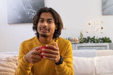 Indian man sipping hot drink on sofa wearing mustard sweater and smartwatch at home holding red mug