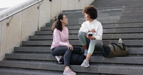Multicultural women sitting on urban steps in athleisure chatting and holding phone