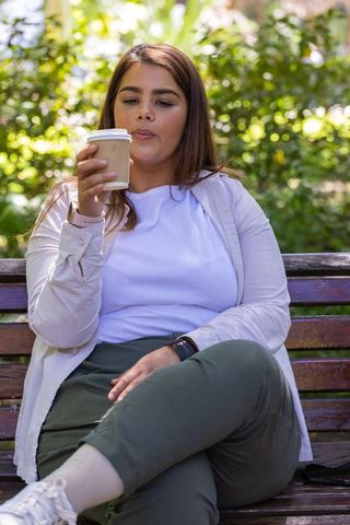 Young woman enjoying takeout coffee on park bench wearing smartwatch, relaxed lifestyle