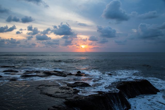 Dramatic scene of sunset over ocean with waves crashing against rocky coastline. Clouds paint vivid patterns across sky as sun dips below horizon. Ideal for portraying natural beauty, serene seascapes, travel destinations, or backgrounds for websites and nature-themed presentations. Evokes calmness and grandeur of nature.