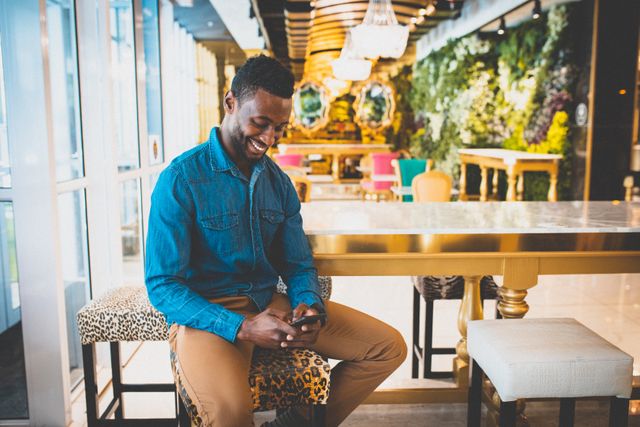 African American man sitting in a stylish, modern cafe, smiling while using his smartphone. He is dressed casually in a denim shirt and brown pants. The cafe has a chic interior with a mix of colorful and patterned furniture. This image can be used for promoting small businesses, technology use, modern lifestyle, or urban living.